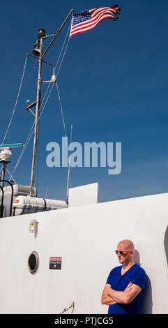 Ein Mann mit einem blauen oberen und Sonnenbrille lehnt sich an eine weiße Wand in eine Fähre unter einem sehr blauen Himmel und die amerikanische Flagge. Stockfoto