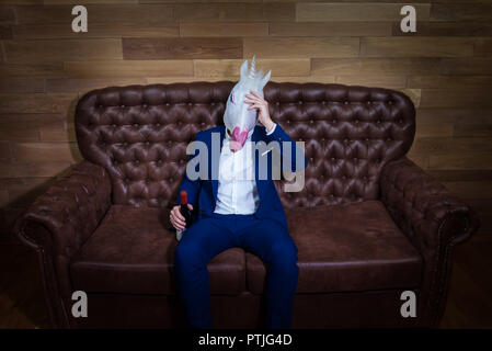 Lustige Einhorn im eleganten Anzug sitzt auf einem Sofa mit einer Flasche Wein. Ungewöhnliche Boss mit Kopfschmerzen zu Hause. Freaky Mann in komischen Maske. Stockfoto