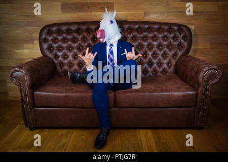 Lustige Einhorn im eleganten Anzug sitzt auf einem Sofa wie ein Chef und zeigt rock Geste. Portrait von ungewöhnlichen Menschen zu Hause. Freaky junge Manager in komischen Maske Stockfoto