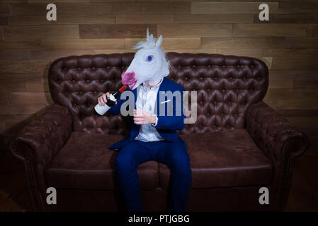 Lustige Einhorn im eleganten Anzug sitzt auf einem Sofa mit einer Flasche Wein. Ungewöhnliche Mann mit Kopfschmerzen zu Hause. Freaky Chef in komischen Maske. Stockfoto