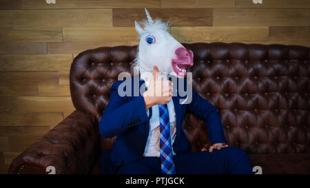 Lustige Einhorn im eleganten Anzug sitzt auf einem Sofa wie ein Chef und zeigt Geste Daumen nach oben. Portrait von ungewöhnlichen Menschen zu Hause. Freaky junge Manager in der Maske Stockfoto