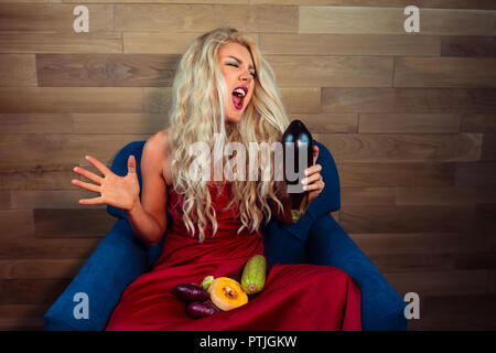 Expressive vegetarische Frau sitzt auf einem Sessel auf den Hintergrund der hölzernen Wand in stilvollen Apartments und singt. Ungewöhnliche Hausfrau mit schönem Haar. Stockfoto