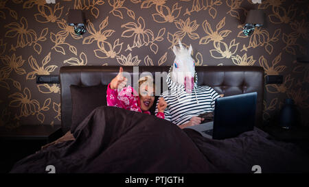 Fröhliches Mädchen zeigt Daumen nach oben Geste und auf dem Bett liegend mit lustigen Freund in komischen Maske. Ungewöhnliche Paar schaut Film auf dem Laptop. Stockfoto