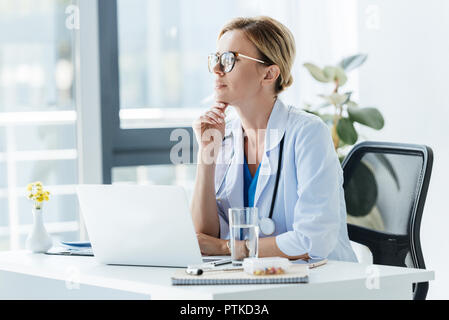 Nachdenklich Ärztin in Brillen am Tisch sitzt mit Laptop im Büro Stockfoto