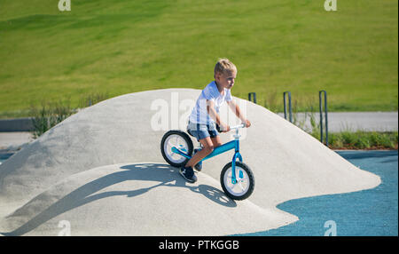 Ein kleiner Junge genießt eine Balance Bike auf künstliche Hügel auf einem Spielplatz in einem Park auf einem sonnigen Tag. Stockfoto