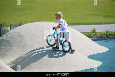 Ein kleiner Junge genießt eine Balance Bike auf künstliche Hügel auf einem Spielplatz in einem Park auf einem sonnigen Tag. Stockfoto