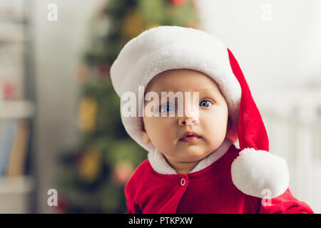 - Nahaufnahme Portrait von entzückenden kleinen Baby in santa hut auf Kamera mit unscharfen Weihnachtsbaum auf Hintergrund Stockfoto