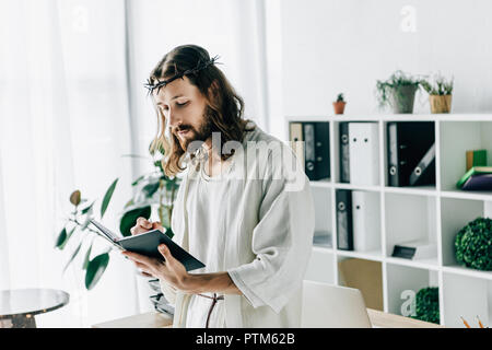 Ernsthafte Jesus mit Dornenkrone und Gewand schreiben in Lehrbuch in der Nähe von Arbeitstisch in modernen Büro Stockfoto