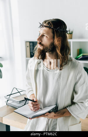 Nachdenklich Jesus mit Dornenkrone und Robe holding Lehrbuch und Wegsehen in modernen Büro Stockfoto