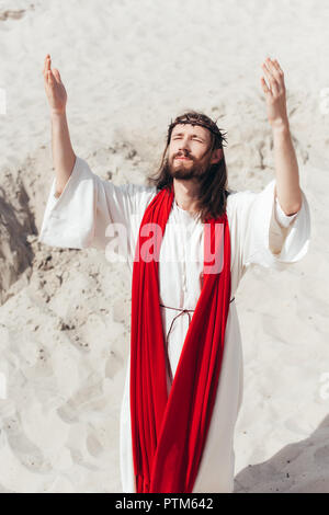 Jesus in Robe, roten Schärpe und Dornenkrone stehend mit erhobenen Händen und betend in der Wüste Stockfoto