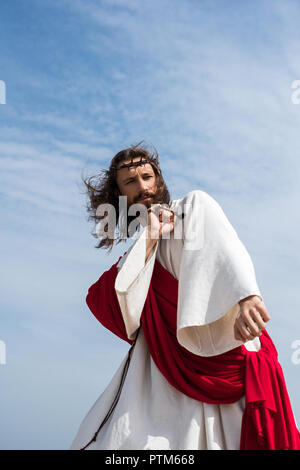 Low Angle View Jesu in Robe, roten Schärpe und Dornenkrone Spaß und Tanzen mit Rosenkranz gegen den blauen Himmel Stockfoto