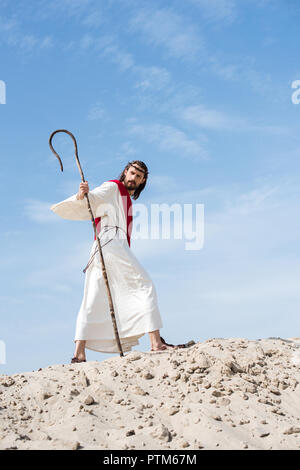 Low Angle View Jesu in Robe, roten Schärpe und Dornenkrone auf sandigen Hügel mit Personal in der Wüste Stockfoto