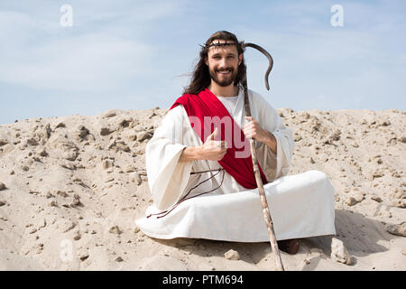 Lächelnd Jesus sitzt im Lotussitz auf Sand, Holding Personal und zeigen Daumen oben in der Wüste Stockfoto
