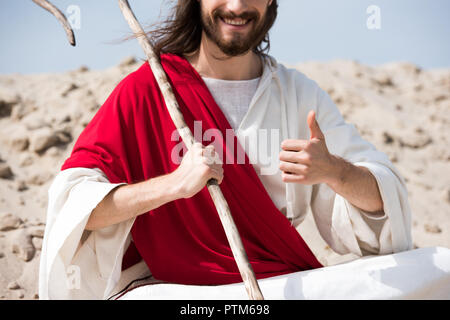 Zugeschnittenes Bild von Jesus im Lotussitz auf Sand sitzen, Holding Personal und zeigen Daumen oben in der Wüste Stockfoto