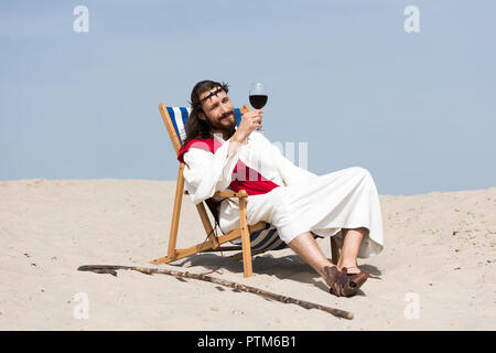 Jesus in Robe und rote Schärpe ruht auf einem Liegestuhl und Blick auf Glas Rotwein in der Wüste Stockfoto