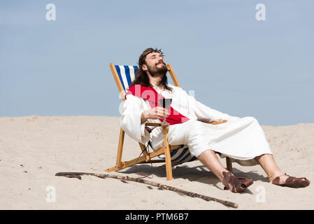 Jesus in Robe und rote Schärpe ruht auf einem Liegestuhl mit einem Glas Rotwein in der Wüste Stockfoto