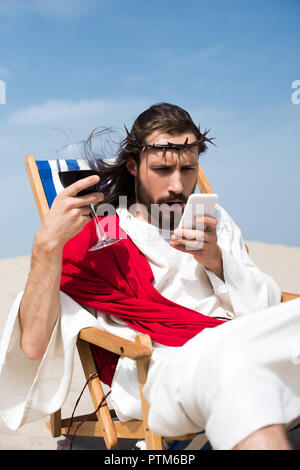 Überrascht Jesus ruht auf einem Liegestuhl mit einem Glas Wein und Smartphone in der Wüste Stockfoto