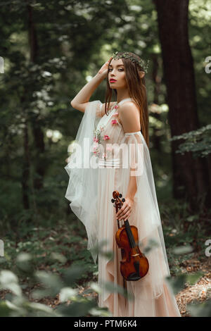 Mystische Elfe im Kleid mit Blüten und Blumen Kranz holding Violine im Wald Stockfoto