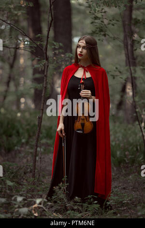 Elegante schöne Frau in schwarzem Kleid und roten Mantel holding Violine im Wald Stockfoto