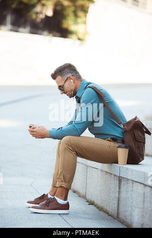 Seitenansicht des Menschen in der Ohrhörer und Sonnenbrille sitzt auf der Treppe und Sie ihr Smartphone Stockfoto