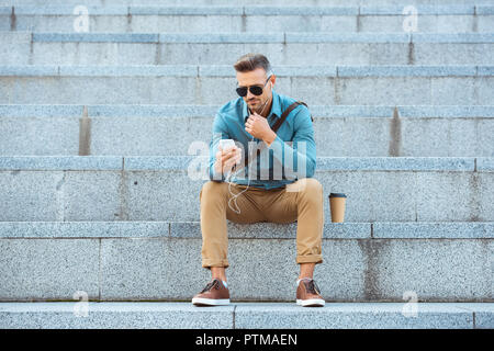 Stilvolle im mittleren Alter Mann in Ohrhörer sitzen auf der Treppe und Sie ihr Smartphone Stockfoto