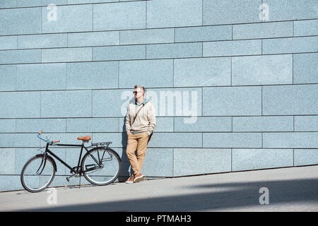 Gut aussehender Mann mittleren Alters in Sonnenbrille mit Händen in den Taschen in der Nähe von Rad- und weg schauen auf der Straße Stockfoto