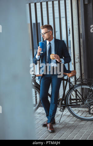 Volle Länge von stattlichen Geschäftsmann holding Hamburger und Trinken aus Papier cup beim Sitzen auf dem Fahrrad und Wegsehen Stockfoto