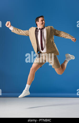 Aufgeregt Mann in Shorts und Anzugjacke springen und weg schauen auf Blau Stockfoto