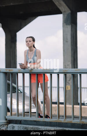 Nachdenklich Sportlerin mit Ohrhörer und Smartphone in der Nähe von Geländer auf Brücke Stockfoto