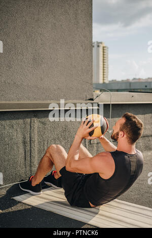 Sportler dabei Sit Ups mit Medizinball auf Yoga Matte auf dem Dach Stockfoto