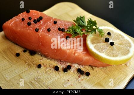 Eine Scheibe roher Lachs gewürzt mit Petersilie, Pfeffer, Salz und Zitrone auf Holz- Häcksler und schwarzen Hintergrund. Seite Vorderansicht Stockfoto
