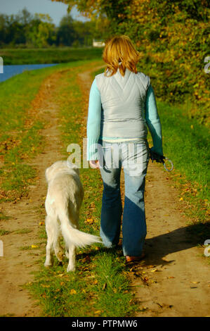 Blonde Frau zu Fuß mit ihrem Golden Retriever entlang des Flusses an einem sonnigen Herbsttag Stockfoto