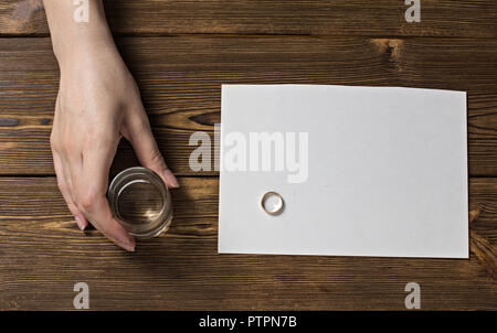 Die Hand einer Frau hält ein Glas mit Alkohol, auf dem Tisch ein Verlobungsring und eine leere Beschriftung Blatt, eine hölzerne Hintergrund Stockfoto