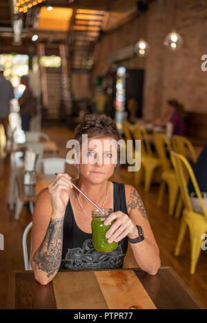 Frau mit Arm Tattoos halten frisch gepressten grünen Saft mit nachhaltigen Metall Stroh am Gelben Backstein Cafe in der Innenstadt von Twin Falls, Idaho. Stockfoto