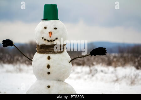 Portrait von Weißen glücklich Schneemann mit orange Karotte Nase, schwarze Steine für die Augen und den grünen Eimer auf den Kopf im Winter, Weihnachten und Stockfoto