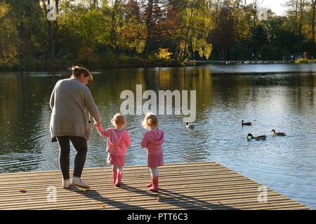 East Renfrewshire, Schottland, Vereinigtes Königreich. Eine Frau und zwei junge Zwillingsmädchen füttern die Enten aus dem Ponton im Rouken Glen Park am Stadtrand von Glasgow. Stockfoto