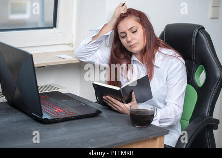 Eine junge Frau, die an einem Schreibtisch sitzen, sie ist umgekippt, betonte, ist sie auf der Suche nach Etwas im Notebook Stockfoto