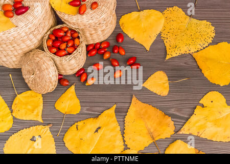 Hagebutten in einen Korb und gelbe Blätter auf einer hölzernen Oberfläche, Konzept der Herbst Hintergrund Stockfoto