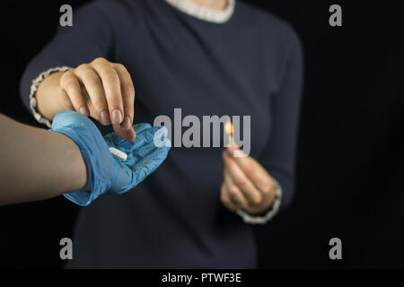 Ein Mädchen auf einem schwarzen Hintergrund hält ein brennendes Streichholz, Schmerzen und Brennen im Magen, die Hand eines Mannes in einem Handschuh hält Ihr eine Pille zu Stockfoto