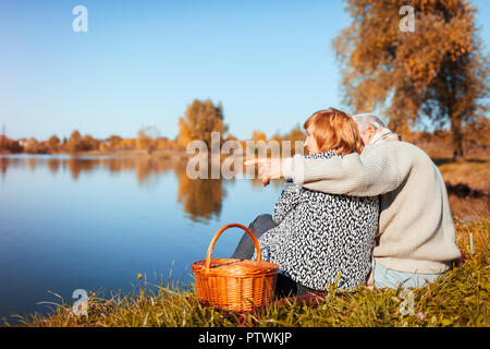 Senior paar Picknick im Herbst See. Glücklich der Mann und die Frau die Natur genießen und umarmen. Mann auf Abstand Stockfoto