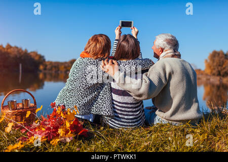 Ältere Eltern unter selfie am Telefon bis Herbst See mit ihrer erwachsenen Tochter. Die Werte der Familie. Menschen mit Picknick Stockfoto