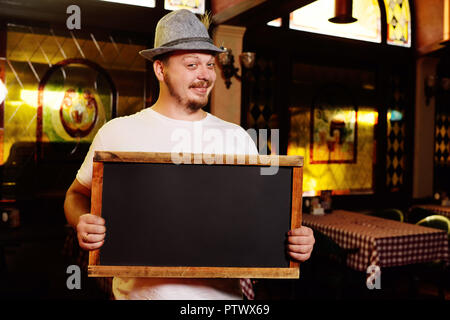 Ein fettes fröhliche Mann in einem bayerischen Hut mit einer Feder bei der Feier der Oktoberfest hält ein Schild oder Tafel in seinen Händen. Für tex, kopieren Stockfoto