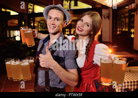 Ein junger Mann und ein Mädchen in der Bayerischen Kleidung Holding eine Menge Becher mit Bier im Pub Hintergrund während der Feier des Oktoberfestes. Stockfoto