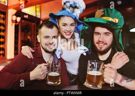 Freunde - Jungen und Mädchen in den Bayerischen hüte Bier trinken auf dem Hintergrund der Bar während der Feier der Oktoberfest oder St. Patrick's Day Stockfoto