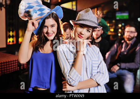 Zwei nette junge Freundinnen in bayerischen Hüte an der Bar Hintergrund lächelt während der Feier der Oktoberfest Stockfoto