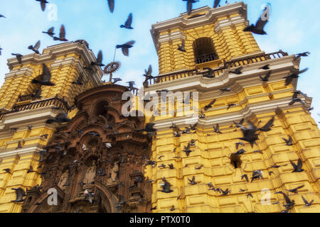 Eine Taubenschar fliegt in Kreisen gegen einen blauen Himmel um die San Francisco Church in Lima, Peru, eine zweifache Kathedrale mit Glockenturm, die gelb gestrichen ist. Stockfoto