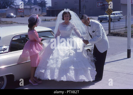 1962, historische, Kalifornien, USA, Sonnenschein, Hochzeit, junge Braut strahlend in ihrem weißen Kleid kommt für Ihren großen Tag mit ihrer Brautjungfer in Rosa und Vater in eine weisse Jacke ihr Begleiter. Stockfoto