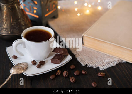 Kleine Tasse schwarzen Kaffee mit Vintage Teelöffel, türkischer Kaffee Topf und Schokolade Stücke auf dunklem Hintergrund mit Buch und Bokeh leuchtet Stockfoto