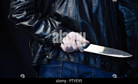 Ein Mann in einer Lederjacke mit einem Messer hinter seinem Rücken, einen schwarzen Hintergrund Stockfoto