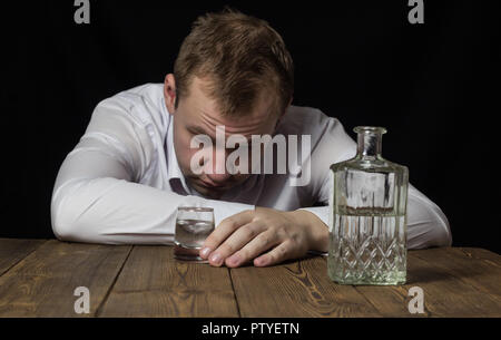 Ein betrunkener Mann in einem weißen Hemd hält ein Glas Alkohol, auf einen hölzernen Tisch ist eine Flasche Alkohol, ein schwarzer Hintergrund Stockfoto